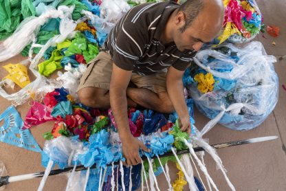 Many sitting on floor weaving together plastic pieces of many colors, surrounded by plastic bags, flags, and sheets of plastic.