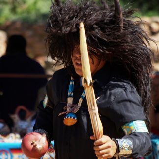 Person with large dark brown hairy head covering with horns. Holding wooden staff and arrows. Wearing black clothing with beaded necklace with shell, armband, and silver and turquoise wrist cuff.