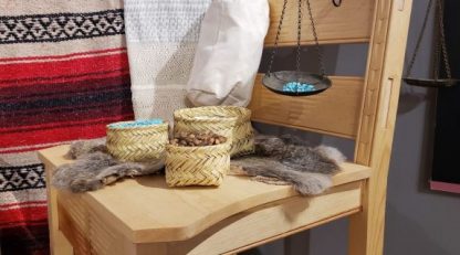 Table with baskets filled with seeds and turquoise stones. Animal fur. Red, black, and white striped blanket, white woven material, metal hanging basket with turquoise hang behind table.