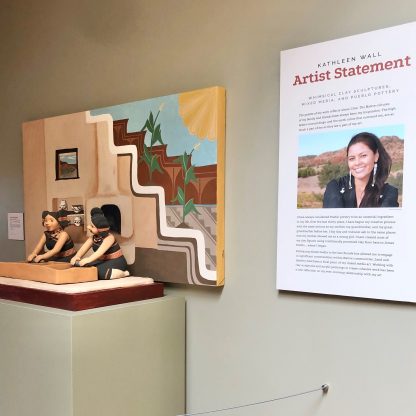 Artist's statement and photo on wall next to painting and sculpture art with two figures working.